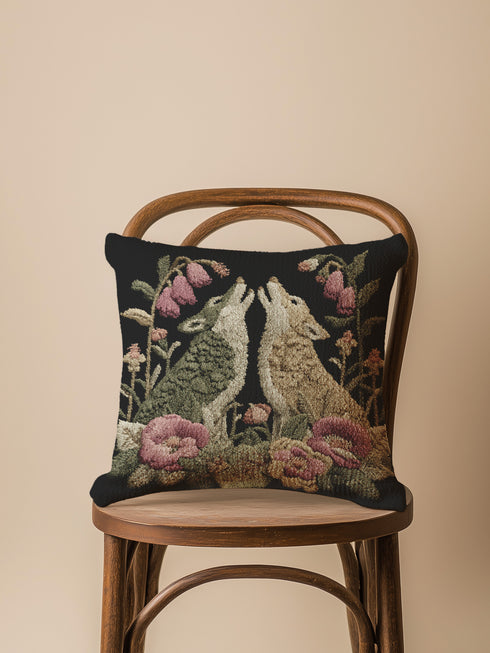 A square woven tapestry pillow with two howling wolves on a black background. The wolves are surrounded by pink and green botanical flowers, creating a vintage, folkloric scene. This decorative cushion is perfect for cottagecore, dark academia, and gothic home decor.